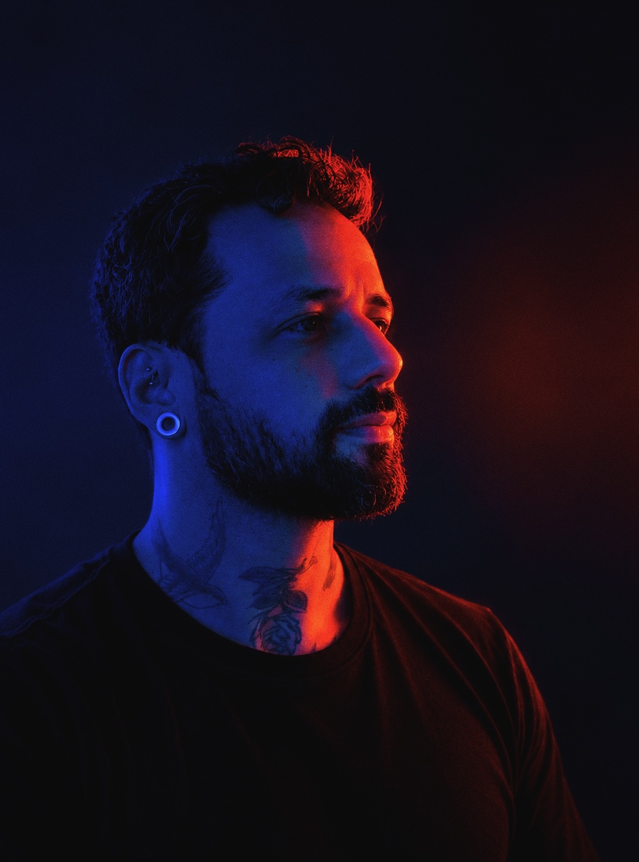 Retrato de Patric Teixeira de perfil, com luz azul de um lado e vermelha do outro: cabelo cacheado, barba, alargador na orelha, tatuagens no pescoço e camiseta preta.
